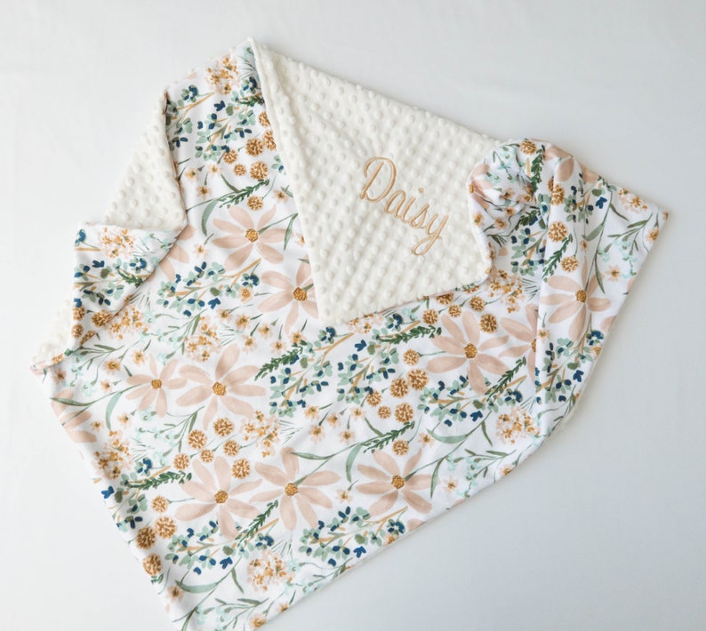 May include: A white and pink floral baby blanket with a white minky backing. The blanket is personalised with the name "Daisy" embroidered in gold thread.
