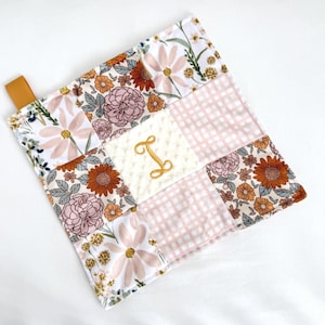 May include: A patchwork baby blanket with floral patterns in pink, orange, and white. The centre square has an embroidered gold letter "I". A tan ribbon is attached to the corner.