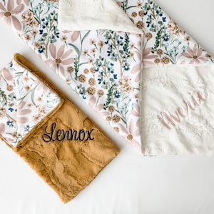 May include: Two personalised baby blankets with floral patterns. One blanket is white with a pink floral pattern and the name "Averie" embroidered on it. The other blanket is brown with the name "Lennox" embroidered on it.