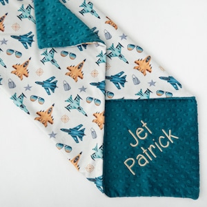Puede incluir: Una manta para bebé azul turquesa y blanca con un nombre bordado personalizado "Jet Patrick" en el lado turquesa. El lado blanco presenta un patrón repetido de aviones azules y naranjas.