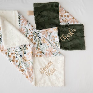 May include: Two personalised baby blankets with floral patterns. One blanket is white with pink flowers and green leaves, and the other is green with white flowers. The white blanket has the name "Lemon Kate" embroidered on it, and the green blanket has the name "Charlie" embroidered on it.