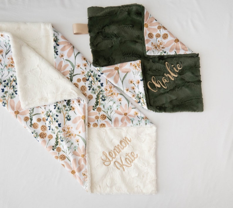 May include: Two personalised baby blankets with floral patterns. One blanket is white with pink flowers and green leaves, and the other is green with white flowers. The white blanket has the name "Lemon Kate" embroidered on it, and the green blanket has the name "Charlie" embroidered on it.