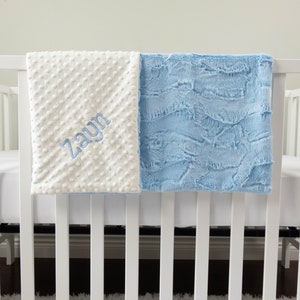 May include: A white and light blue baby blanket with the name "Zayn" embroidered on the white side. The blanket is hanging over a white crib.