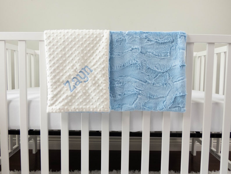 May include: A white and light blue baby blanket with the name "Zayn" embroidered on the white side. The blanket is hanging over a white crib.