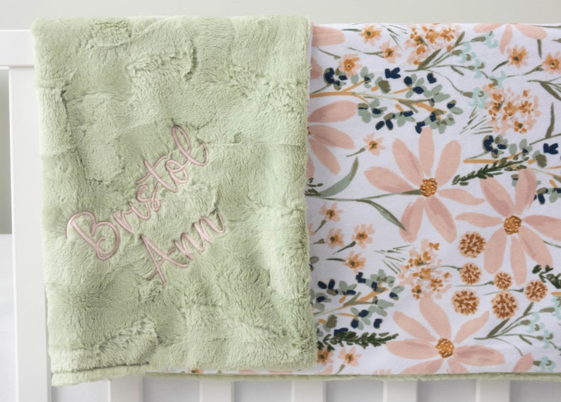 May include: A green plush baby blanket with a floral print on the reverse. The blanket is personalised with the name "Bristol Ann" embroidered in pink thread.