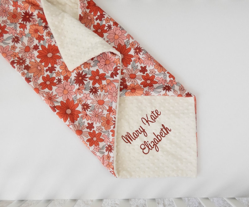 May include: A white and orange floral baby blanket with a soft, white minky backing. The blanket is personalized with the name "Mary Kate Elizabeth" embroidered in red.