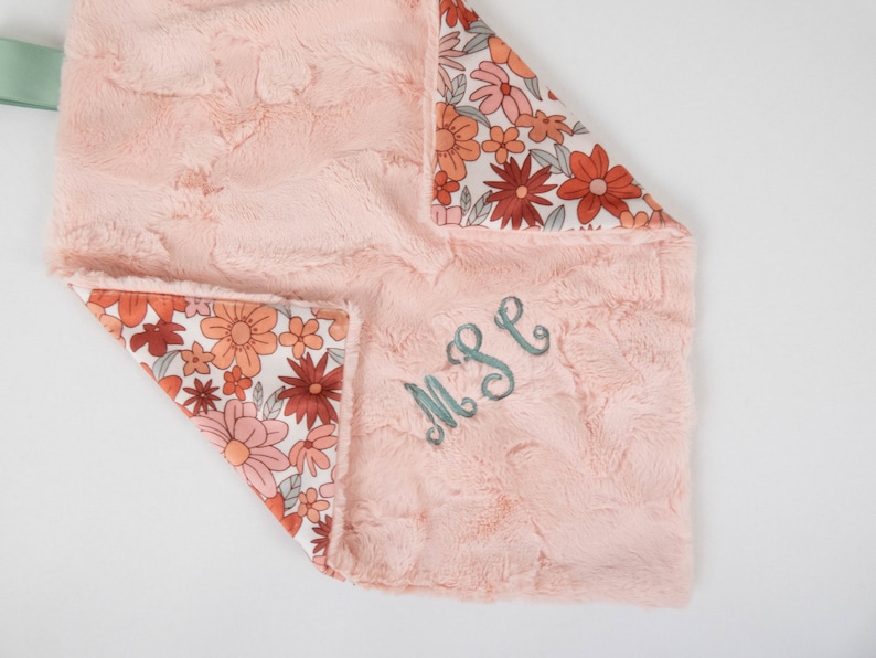 May include: A pink and white baby blanket with a floral pattern on one side and a soft, fuzzy texture on the other. The blanket is personalized with the monogram "MPE" in teal thread.