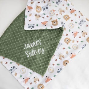 May include: A baby blanket with a forest animal print in orange, brown, and gray on a white background. The blanket has a green, textured section with the embroidered name "James Sidney".