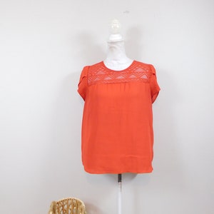 Può includere: Una camicetta arancione-rossa con maniche corte e un pannello in pizzo decorativo sulla parte superiore del petto. La camicetta è esposta su un manichino bianco su uno sfondo bianco. Un cesto di vimini è in primo piano.