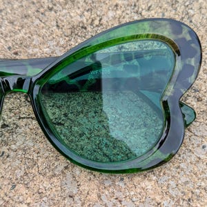 K&ouml;nnte beinhalten: Gr&uuml;ne, schmetterlingsf&ouml;rmige Sonnenbrille mit dunkelgr&uuml;nem Rahmen und gr&uuml;nen Gl&auml;sern. Die Sonnenbrille hat den Text "WF 1205" auf der Innenseite der rechten Linse.