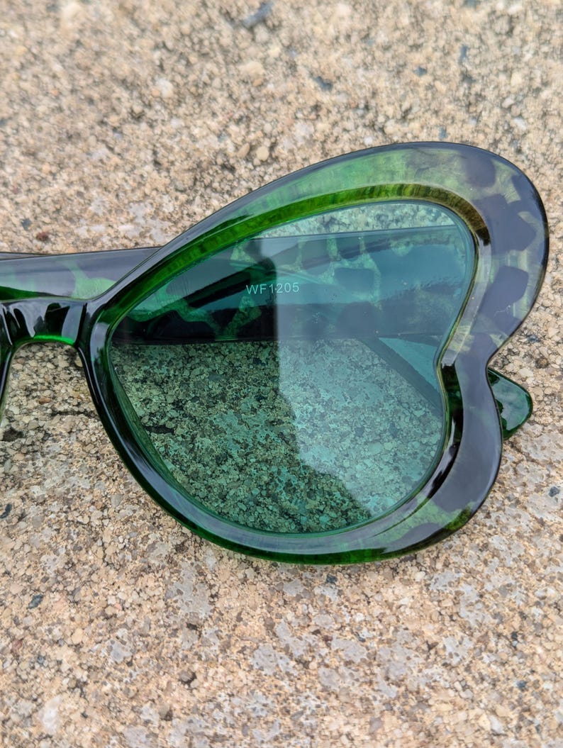 K&ouml;nnte beinhalten: Gr&uuml;ne, schmetterlingsf&ouml;rmige Sonnenbrille mit dunkelgr&uuml;nem Rahmen und gr&uuml;nen Gl&auml;sern. Die Sonnenbrille hat den Text "WF 1205" auf der Innenseite der rechten Linse.
