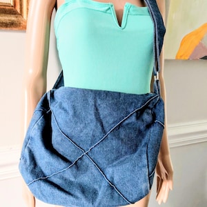 May include: A medium-wash blue denim crossbody bag featuring a quilted diamond pattern created by visible stitching across the front panel. The bag has a wide shoulder strap and a slouchy, unstructured shape, worn over a light mint green ribbed tank top.