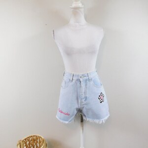 May include: Light blue denim shorts with frayed edges, displayed on a mannequin. The shorts feature the word "Optimistic" in pink script on the front and a flower patch with a cow print design.