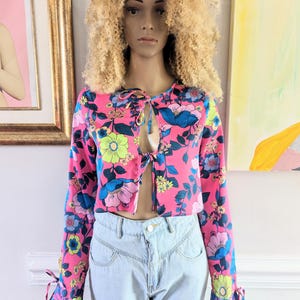 May include: A pink, blue, and yellow floral print cropped top with long sleeves and tie detail at the neckline. The sleeves have tie details at the cuffs.