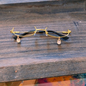 Puede incluir: Gafas con montura dorada y patillas negras con colgantes en forma de lágrima. Las gafas tienen un diseño delicado y minimalista, con una fina montura dorada y una estética sutil y elegante. Los colgantes de lágrima añaden un toque de sofisticación.