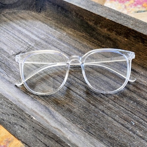 May include: Clear, round-framed eyeglasses with a transparent plastic frame. The glasses are set against a dark, textured wooden surface. The lenses are clear, and the overall design is minimalist and modern.