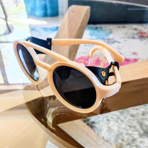 May include: Cream-colored sunglasses with black lenses. The sunglasses have a unique design with a thick frame and a black strap detail. The lenses are oval-shaped, and the frame is resting on a reflective surface.
