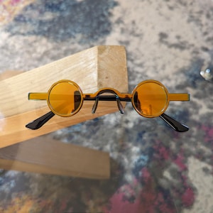 May include: A pair of round, amber-coloured sunglasses with black frames. The sunglasses are sitting on a wooden surface.