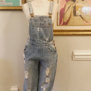 May include: Light blue denim overalls with distressed details and a single front pocket. The overalls are on a white mannequin.