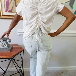 May include: A white, short-sleeved shirt with a cinched back detail. The shirt is worn with light blue jeans.