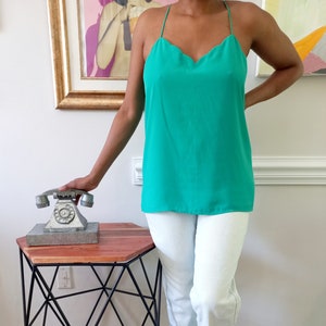 May include: A bright green camisole top with a scalloped neckline and spaghetti straps. The top is paired with white pants. A vintage silver telephone sits on a small, hexagonal table with a black metal frame.