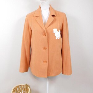 May include: A peach-colored blazer with a three-button closure and a white poodle applique on the front. The blazer is displayed on a mannequin and has a classic lapel design. A wicker basket is in the foreground.