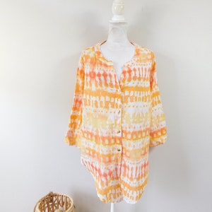 May include: A long-sleeved shirt in a tie-dye pattern of orange, yellow, and white, displayed on a mannequin. The shirt has a mandarin collar and gold-coloured buttons. A woven basket is in the foreground. The shirt is made of a light fabric.