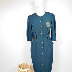 Puede incluir: Vestido color verde azulado con botones dorados y un parche bordado decorativo en el pecho. El vestido tiene mangas tres cuartos y una botonadura frontal. El vestido se exhibe en un maniquí blanco.