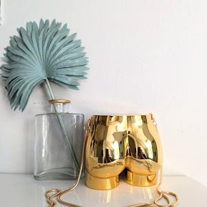 May include: A gold-coloured novelty handbag shaped like a pair of buttocks. The purse has a gold chain strap and is displayed on a white surface. A clear glass vase with a blue-green decorative leaf is in the background.