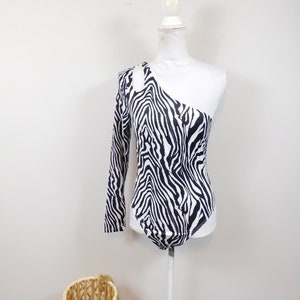 Pode incluir: Um body de uma só manga com estampa de zebra em preto e branco. O body tem um detalhe de corte no ombro.