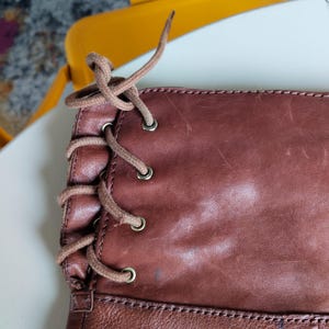 Pu&ograve; includere: Primo piano di uno stivale in pelle marrone con design allacciato. Lo stivale presenta un ricco colore marrone-rossastro con texture e usura visibili. I lacci sono di un marrone pi&ugrave; chiaro, in contrasto con la pelle pi&ugrave; scura. Gli occhielli sono metallici.