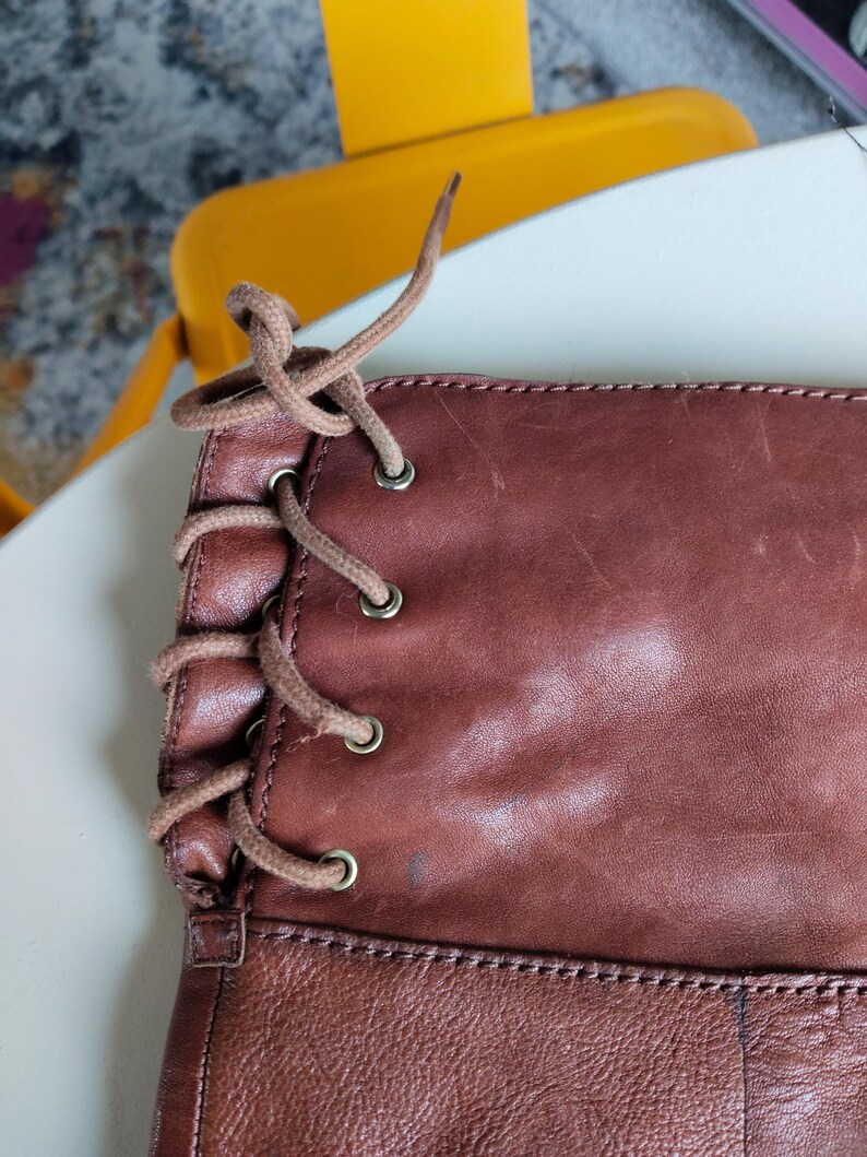 Pu&ograve; includere: Primo piano di uno stivale in pelle marrone con design allacciato. Lo stivale presenta un ricco colore marrone-rossastro con texture e usura visibili. I lacci sono di un marrone pi&ugrave; chiaro, in contrasto con la pelle pi&ugrave; scura. Gli occhielli sono metallici.