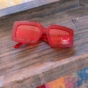 Puede incluir: Gafas de sol rojas con montura cuadrada y lentes rojas. Las gafas de sol tienen una montura gruesa y un diseño clásico. Las lentes tienen protección UV. Las gafas de sol están sobre una superficie de madera.