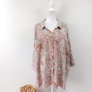 May include: A button-down shirt with a paisley pattern in shades of olive green, red, and pink. The long-sleeved shirt is displayed on a white mannequin. A woven basket is in the foreground.