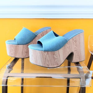 May include: A pair of blue denim platform sandals with wooden-effect heels. The sandals have a thick sole and a wide strap. The heels are approximately 10 cm high.