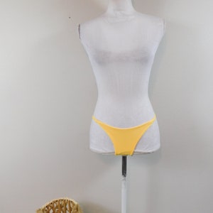 Könnte beinhalten: Ein gelber Bikini-Slip an einem weißen Mannequin. Der Bikini-Slip ist einfarbig und schlicht mit einem hohen Beinausschnitt.