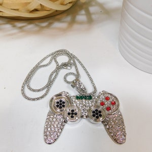 Könnte beinhalten: Silberfarbene Anhängerkette mit einem Strass-besetzten Videospiel-Controller-Design. Der Controller hat schwarze und rote Knöpfe und die Buchstaben "MAS" in Grün. Die Halskette ist ebenfalls silberfarben.