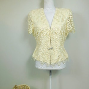 May include: A cream-colored lace top with a V-neckline and short sleeves. The top has a scalloped hem and a button closure at the front. The top is on a white mannequin.