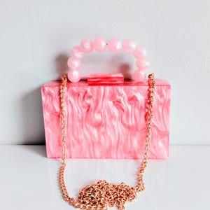 Puede incluir: Un bolso de mano tipo caja rectangular con un acabado acrílico jaspeado en rosa y blanco. El bolso incluye un asa superior hecha de cuentas rosas redondas y translúcidas, una correa de cadena metálica dorada desmontable y un cierre rectangular.