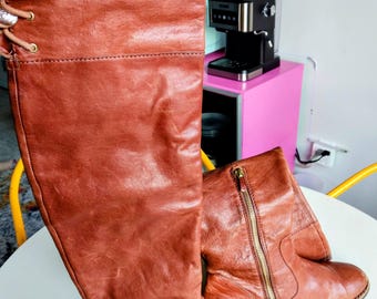 May include: A pair of brown leather knee-high boots. One boot is upright, showing the full height and lace-up detail, while the other is lying down, revealing a side zipper. The boots have a worn, textured appearance.