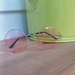 Puede incluir: Un par de gafas de sol redondas de forma octogonal con montura dorada y lentes rosas. Las gafas de sol están sobre una superficie de madera.