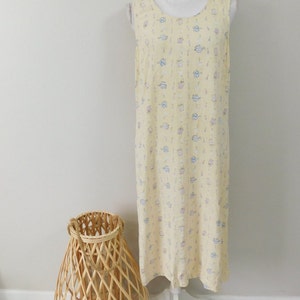 Puede incluir: Un vestido sin mangas de color amarillo claro con un estampado de pequeñas tazas de té y teteras azules y moradas. El vestido se exhibe en un maniquí blanco. Una cesta tejida y una pequeña planta en maceta están en primer plano.