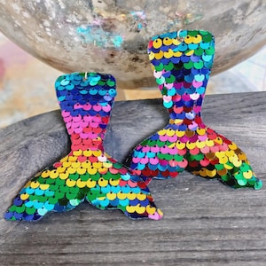 May include: A pair of rainbow mermaid tail earrings made of iridescent sequins. The earrings are a fun and festive accessory for any occasion.