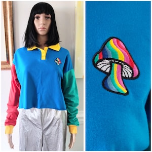 May include: A colour-blocked long-sleeve shirt with a blue body, red left sleeve, and green right sleeve. The shirt has a yellow collar and cuffs. A colourful mushroom patch is on the chest.