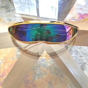 Könnte beinhalten: Goldgerahmte Sonnenbrille mit futuristischem Design. Die Gläser haben eine reflektierende, irisierende Beschichtung, die ein Farbspektrum zeigt. Die Sonnenbrille liegt auf einer transparenten Oberfläche und reflektiert Licht und Farben.