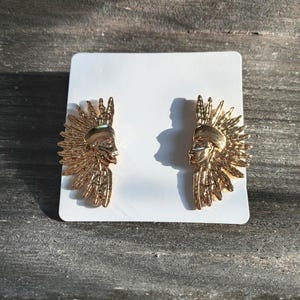 May include: A pair of gold-colored earrings featuring a Native American chief design. Each earring has a profile of a face with a feathered headdress. The earrings are displayed on a white card against a weathered wood background.