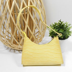 Puede incluir: Un pequeño bolso amarillo con un diseño texturizado en relieve de cocodrilo. El bolso tiene una correa de hombro corta y una cremallera superior.