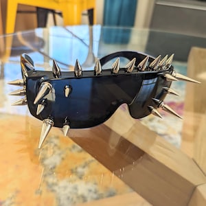 May include: Black sunglasses with silver spikes around the frame. The sunglasses have a futuristic, cyberpunk aesthetic.