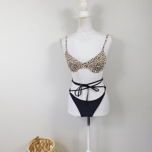 Op de afbeelding: Een zwarte en bruine bikini top met luipaardprint en een zwarte string bikini broekje. Het bikini broekje heeft een hoge beenuitsnijding en knoopt aan de zijkanten.