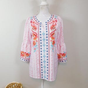 May include: A white, pink, and blue embroidered tunic top with a floral pattern. The top has a V-neck and 3/4 sleeves.